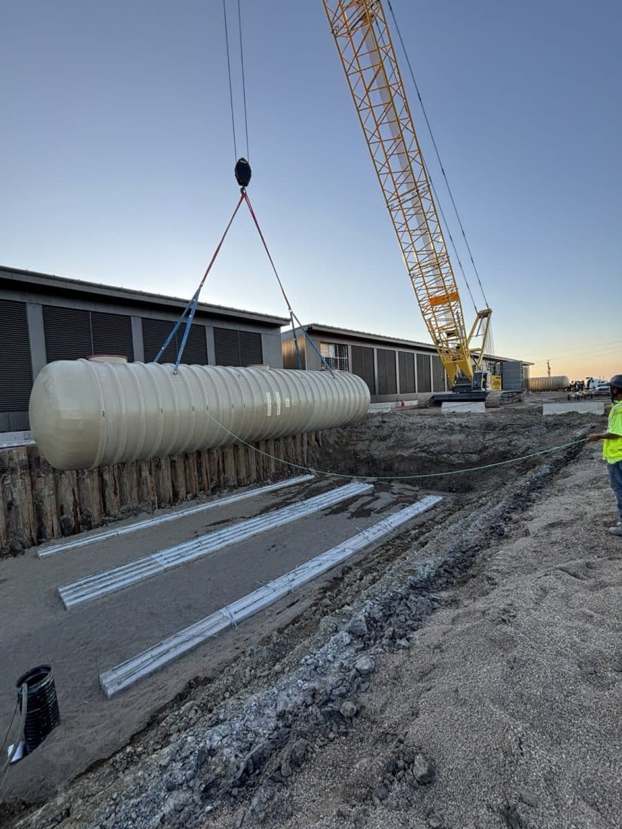 Crawler crane lowering a fiberglass fuel storage tank into an excavation at dusk